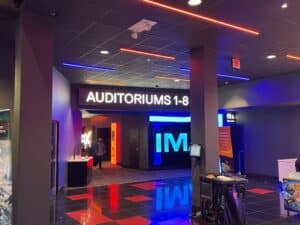 a Modern Frederick Movie Theatre Lobby with Glowing Signs a Large auditoriums 1 8 Sign Hangs from the Ceiling Entrances Below Neon Lights a Ticket Kiosk and Movie Posters Line the Walls As Two People Walk Inside