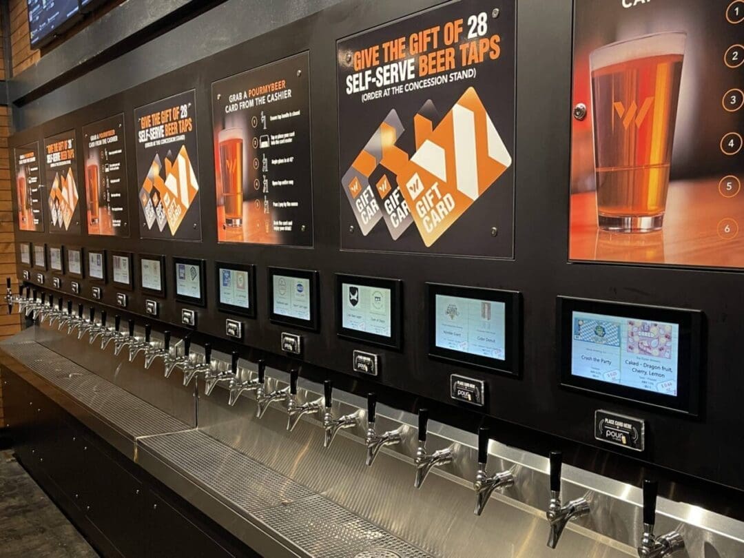 a Row of 28 Self Serve Beer Taps is Mounted on a Black Wall in This Frederick Movie Theatre Each with a Digital Display Above Large Signs Promote Gift Cards and the Self Serve Feature with a Glass of Beer Shown on the Signs