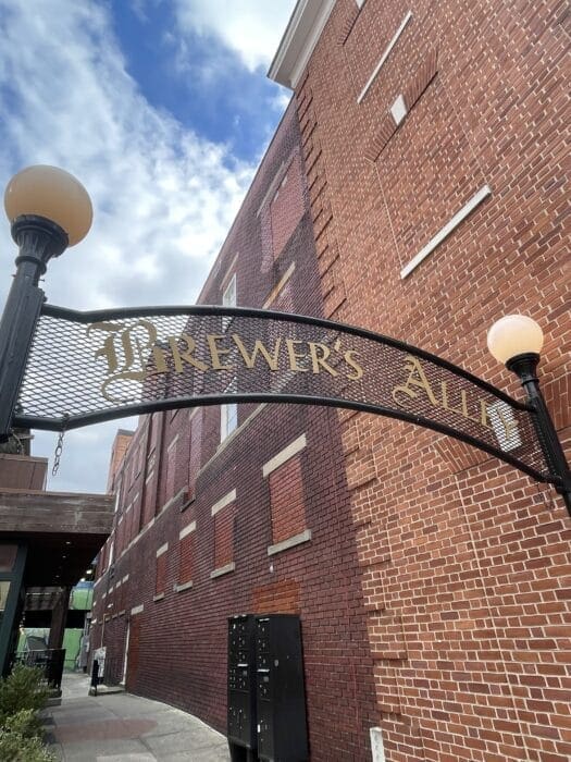 A decorative black iron archway with gold letters reading Brewer’s Alley, one of the best bars in Frederick, stands between two lampposts in front of a tall red brick building. The sky is partly cloudy, and a sidewalk runs alongside the building.