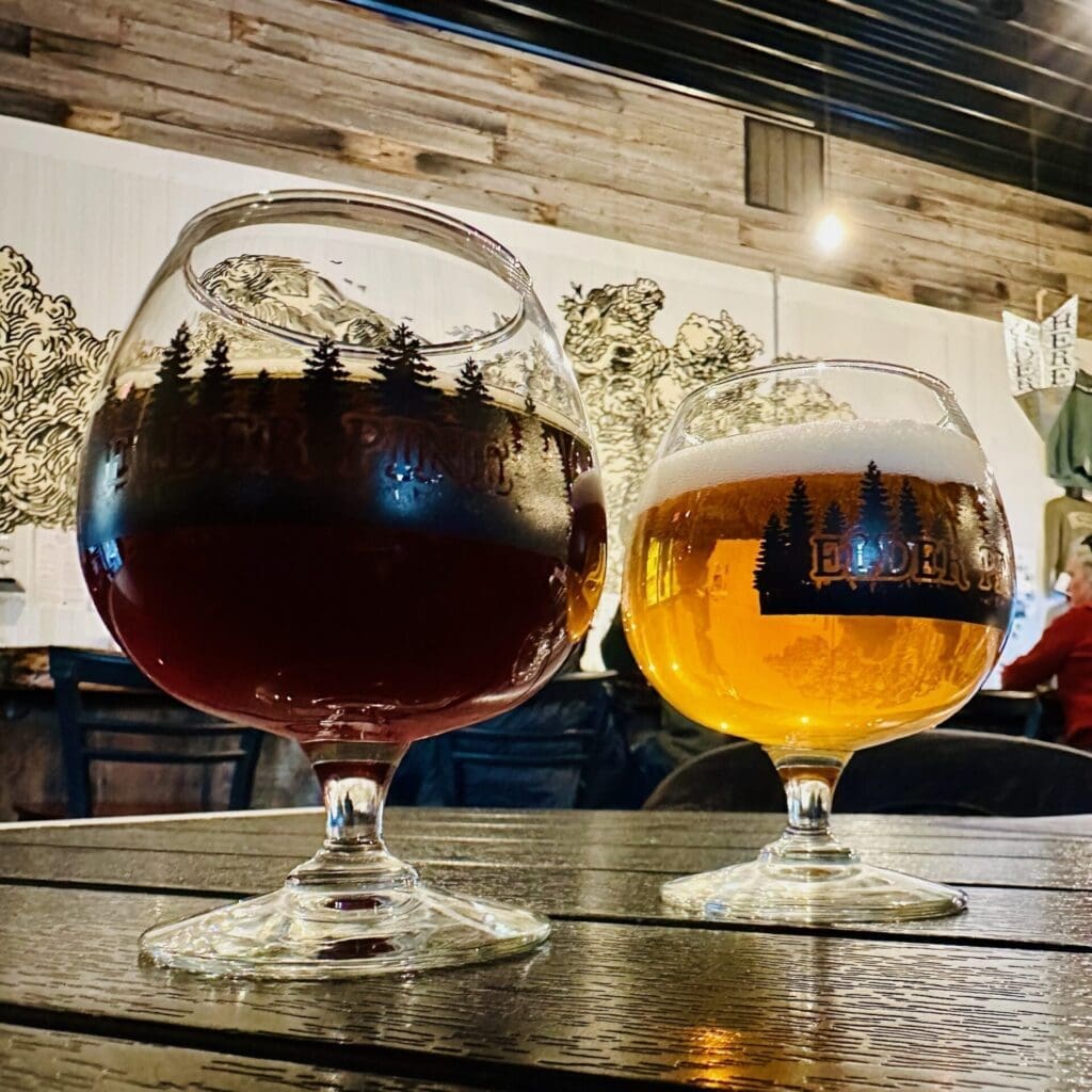 Two large snifter glasses of beer from the best breweries in Frederick MD sit on a wooden table. One is dark brown with foam, the other golden and clear. Both feature forest silhouettes, set against the cozy backdrop of a local bar.
