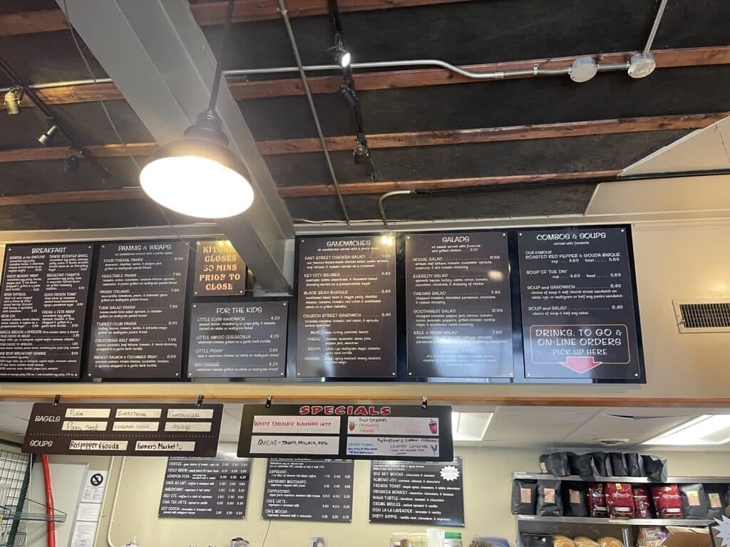 a Deli Counter Menu Hangs Above a Counter Listing Breakfast Sandwiches Salads Combos and Specials in White Text on Black Boards Perfect for a Quick Bite at a Coffee Shop in Frederick Md Fresh Bread Bags Are Stacked on Shelves Below