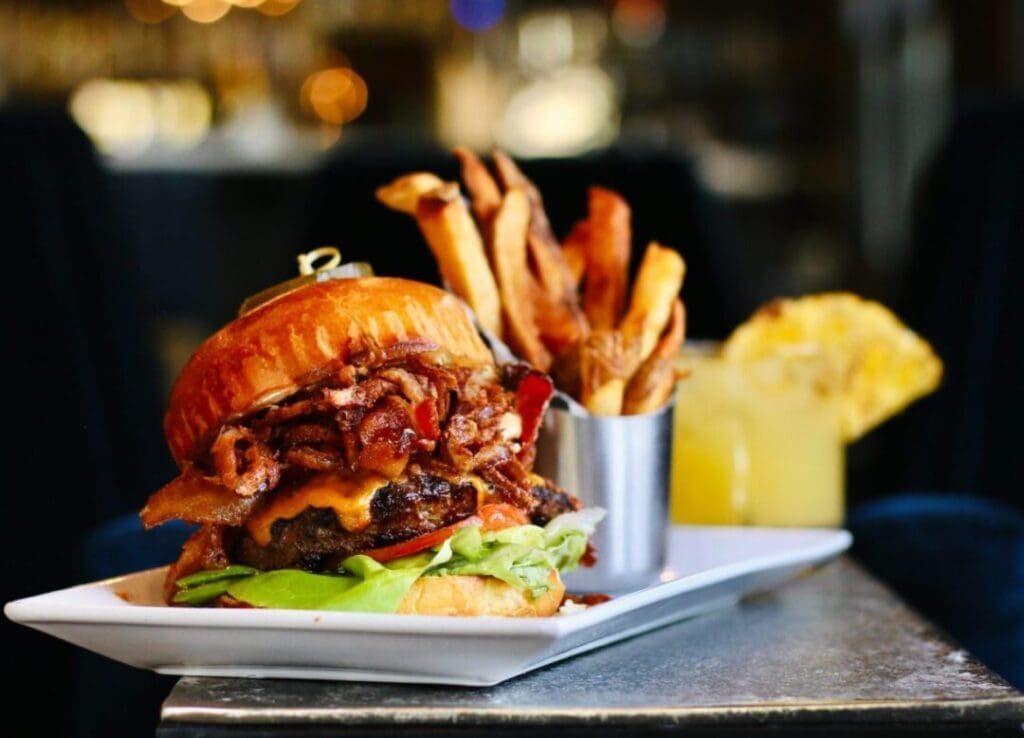 a Cheeseburger Stacked with Crispy Bacon Lettuce and Tomato is Served on a White Plate Beside Golden French Fries in the Background a Yellow Drink with a Pineapple Garnish Hints at the Best Burger Frederick Md Has to Offer