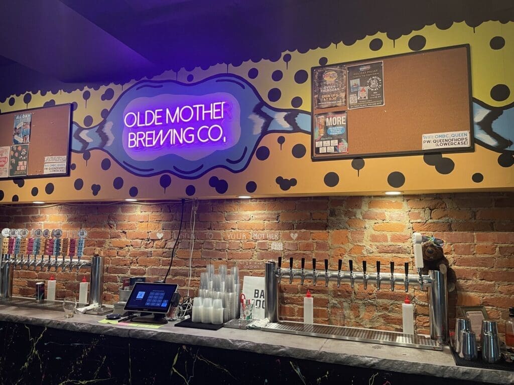 Bar counter at Olde Mother Brewing Co., one of the best breweries in Frederick MD, with a neon sign on a cheetah-print wall above brick, showing beer taps, drink cups, a cash register, and community boards. The bar is brightly lit with modern and rustic elements.