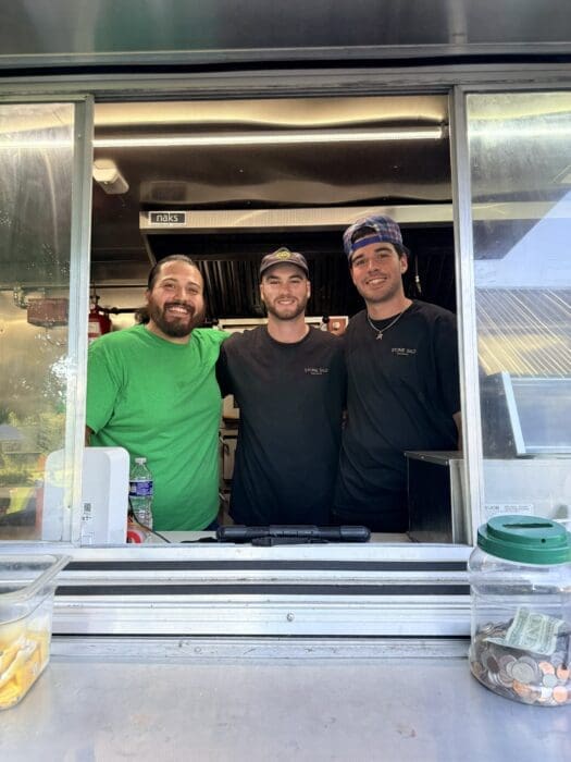 Three Smiling Staff Members Stand Together in a Food Truck Window Wearing Casual Shirts green Black and Black with a Cap