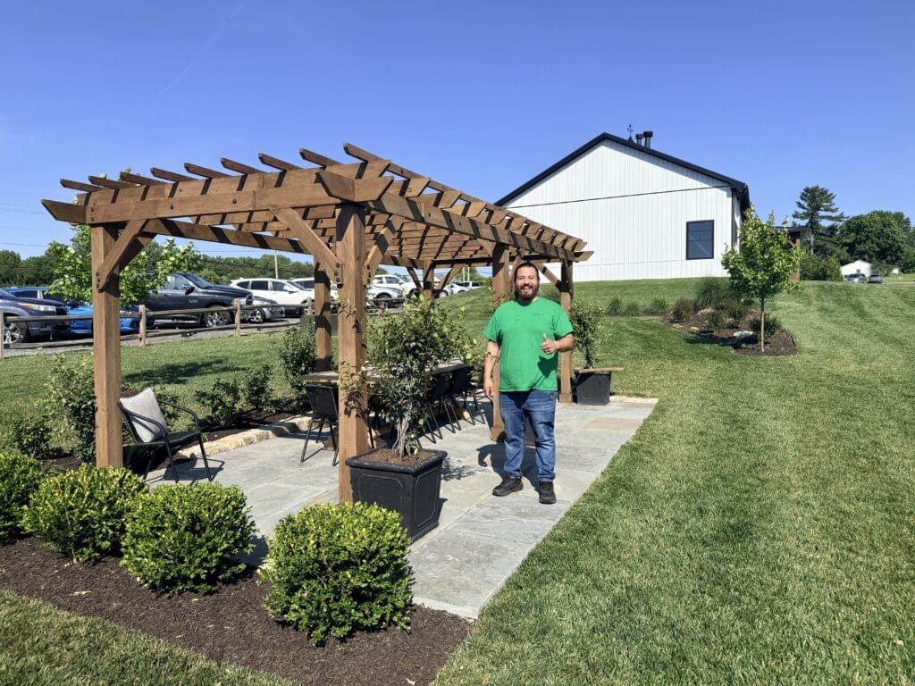 Man in a Bright Green Shirt Stands Under a Large Wooden Pergola Giving a Thumbs Up with a Modern White Building and Parked Cars in the Background on a Sunny Day