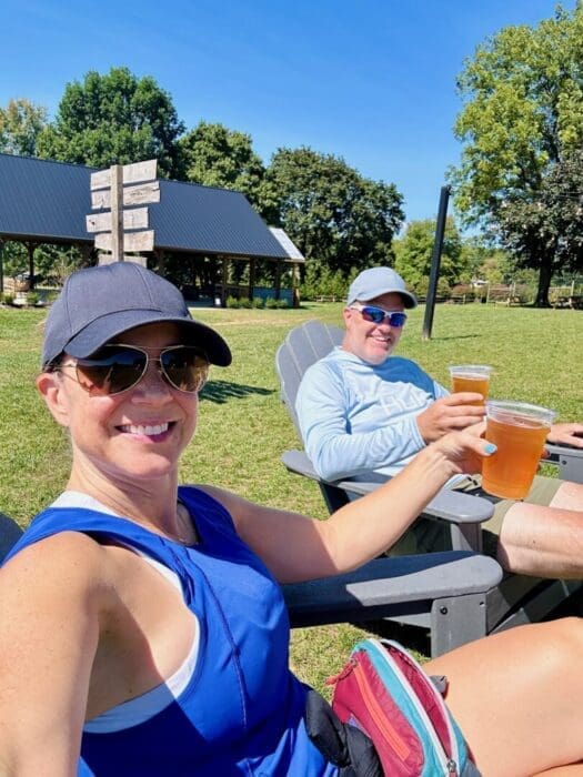 A woman and man sit outdoors in Adirondack chairs at one of the best breweries in Frederick MD, smiling and holding up plastic cups of beer. Both wear sunglasses, hats, and casual summer clothes, framed by green grass, trees, and a pavilion under clear blue skies.