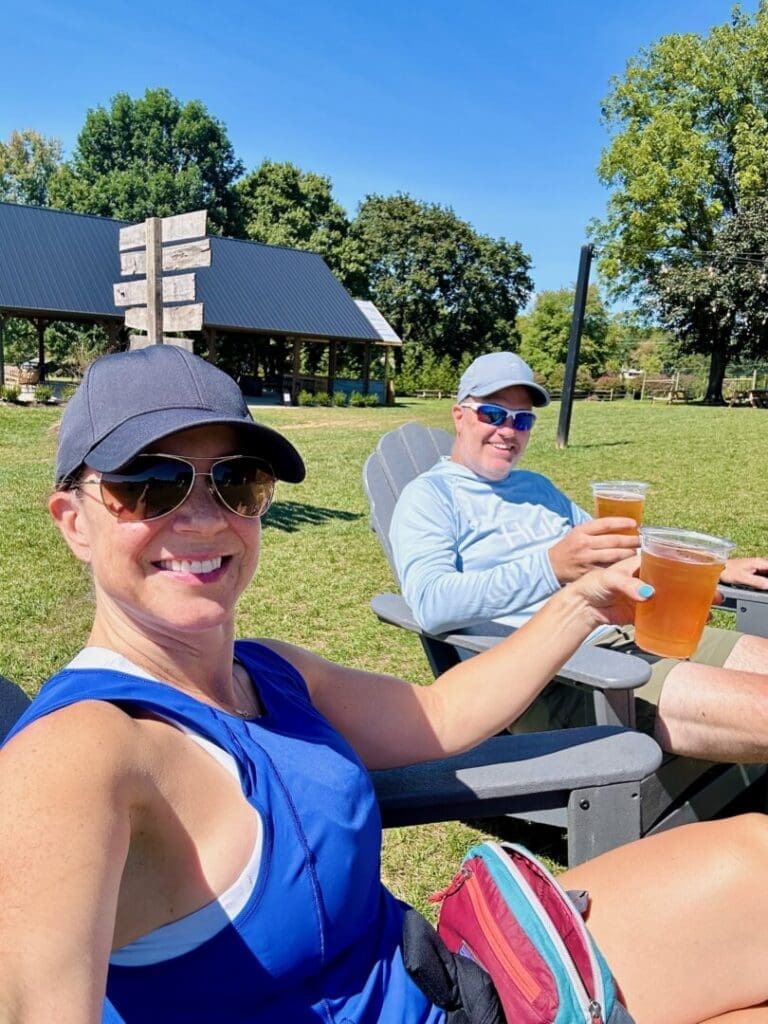 A woman and man sit outdoors in Adirondack chairs at one of the best breweries in Frederick MD, smiling and holding up plastic cups of beer. Both wear sunglasses, hats, and casual summer clothes, framed by green grass, trees, and a pavilion under clear blue skies.