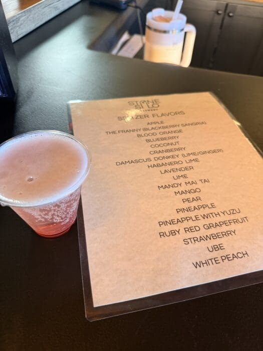 Pink Smoothie in a Plastic Cup Sits on a Dark Counter Beside a Beige Flavor Menu a Blender Pitcher is Visible in the Background