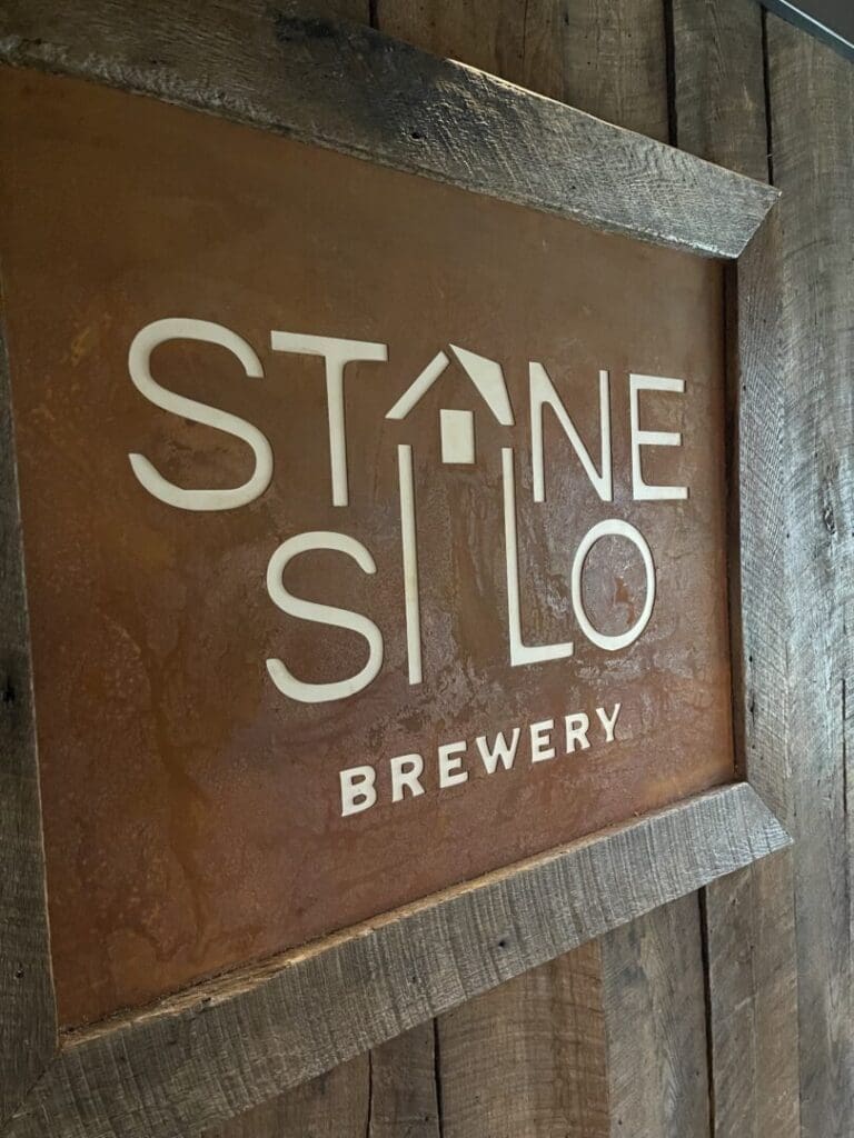 A rustic wooden sign displays the text Stone Silo Brewery in white, with the A in Stone styled as a roof with a small chimney to resemble a house. Set against a weathered, rusty orange background and rough wooden frame, it captures New Market MD charm.