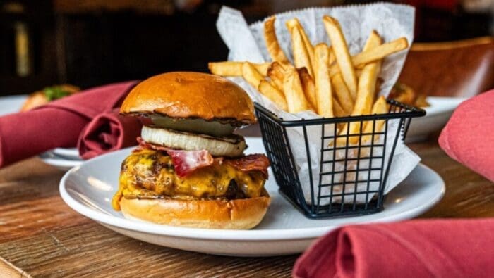 a Cheeseburger with Melted Cheddar Bacon and a Thick Slice of Onion on a Shiny Bun Sits on a White Plate Beside Golden French Friesclassic Comfort Food Youll Find at Top Restaurants in Frederick Md Red Napkins Brighten the Wooden Table