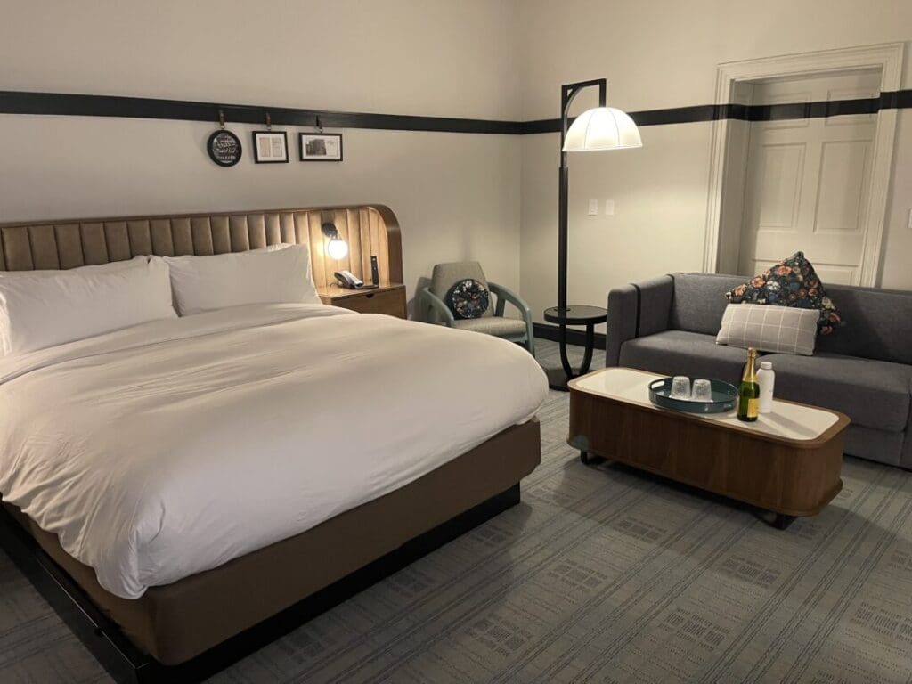 A cozy hotel room in one of the top hotels in Frederick MD, featuring a large bed with a gray headboard, white bedding, a gray armchair, sofa with patterned pillows, wooden coffee table with drinks, wall art above the bed, and a floor lamp.