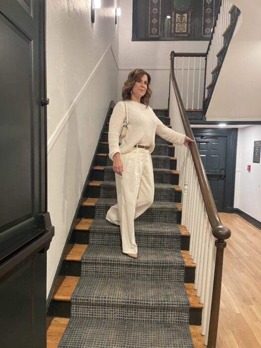 A woman with wavy brown hair, wearing a cream sweater and wide-leg white pants, stands midway on a carpeted staircase indoors—her style echoing the modern, clean interior design found in boutique hotels Frederick MD is known for.