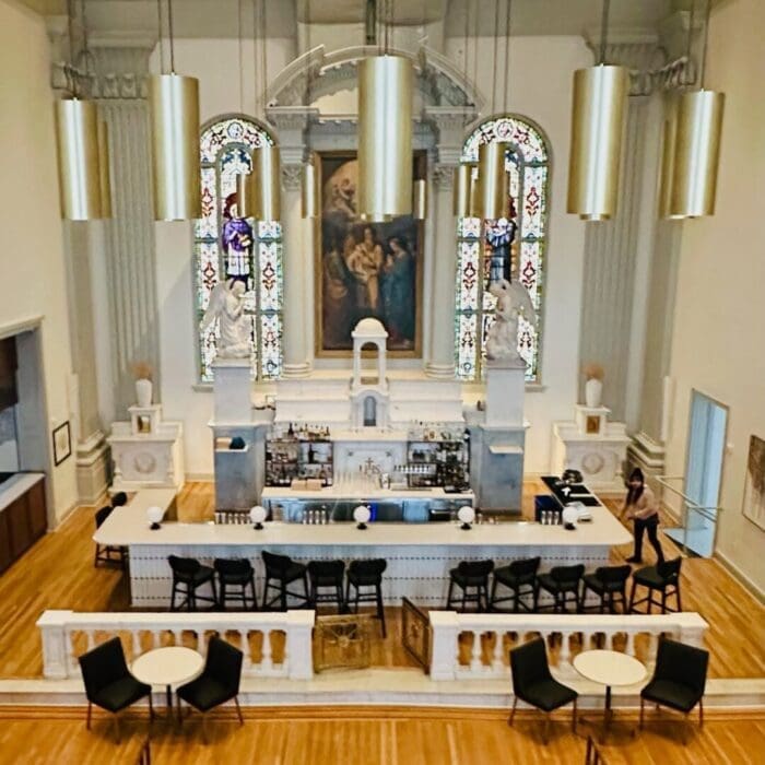 A stylish bar set inside a former church, this unique gem among restaurants in Frederick MD features high stained-glass windows, neoclassical columns, gold pendant lights, a marble bar lined with bottles, black barstools, and round tables on a wooden floor.
