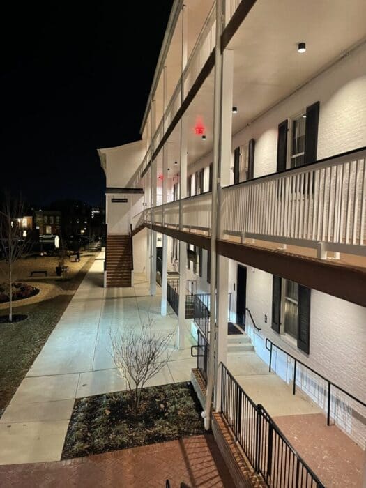 A two-story white brick building at night features a lit walkway, black shutters on windows, and white railings. External stairs lead to the upper floor. Leafless trees and landscaped areas border the walkway at Visitation Hotel Frederick MD.