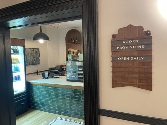 a Small Shop Inside Visitation Hotel Frederick Md Features a Green Tiled Counter Fridge with Drinks Shelves of Items and a Register a Wooden Sign Reads Acorn Provisions Open Daily the Space is Clean Modern and Well Lit