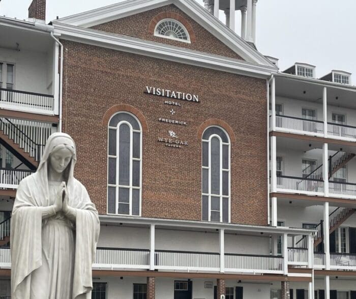 A white statue of the Virgin Mary stands with hands in prayer before a brick building labeled “Visitation Hotel Frederick” in Frederick, Maryland. The building has tall arched windows, white trim, and a small rooftop tower under an overcast sky.