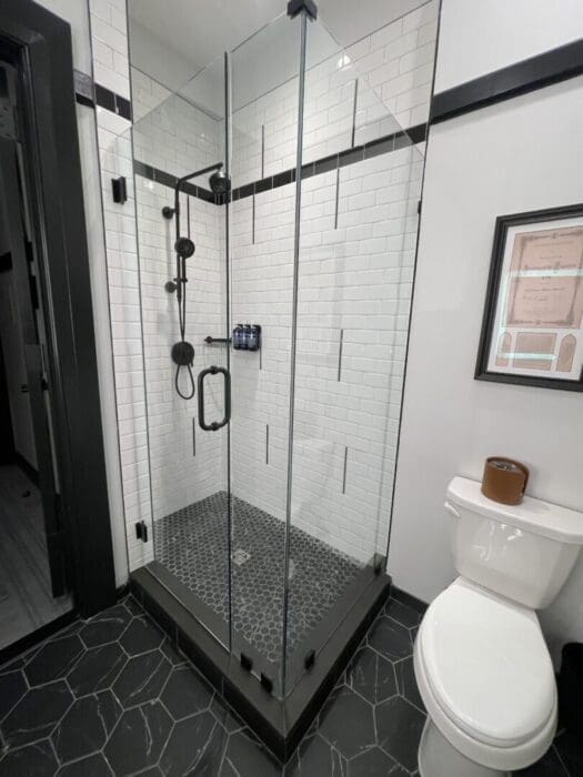 a Modern Bathroom at Visitation Hotel Frederick Md Features a Glass Shower with White Subway Tile Walls Black Fixtures and Hexagonal Black Tile Floor Outside the Shower is a White Toilet Black and White Walls a Framed Picture and Sleek Finishes Throughout