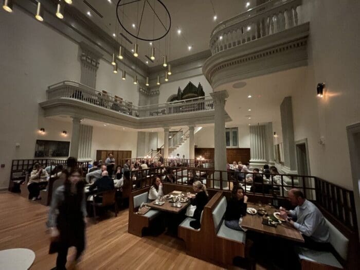 A spacious, elegant restaurant with high ceilings, chandeliers, and a balcony—perfect for guests from boutique hotels Frederick MD. Soft lighting, wooden floors, and classical details create a warm atmosphere as diners enjoy meals and attentive service.