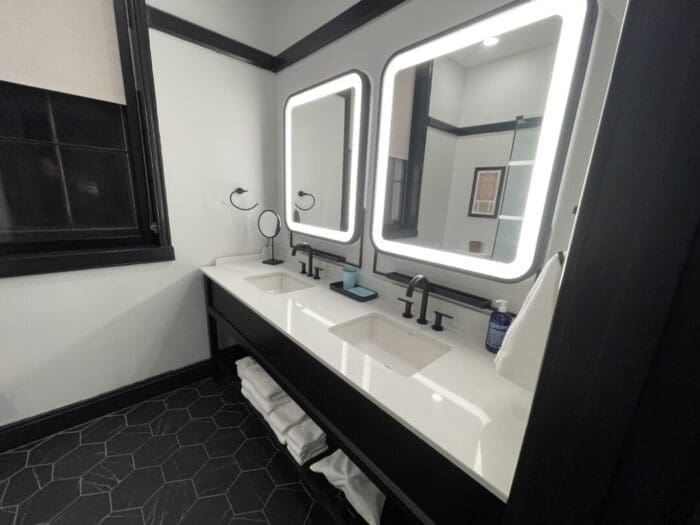 a Modern Bathroom at Visitation Hotel Frederick Md Features Two Rectangular Sinks with Backlit Mirrors Black Faucets and a Black Countertop Shelves Hold White Towels Beneath While Black Hexagonal Floor Tiles and Black trimmed Windows Complete the Sleek Look