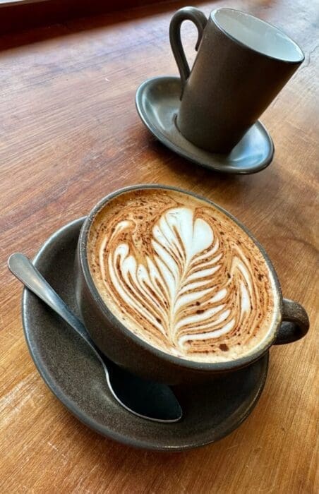 A cup of latte with intricate leaf-shaped art sits on a brown saucer with a spoon, on a wooden table. In the background, an empty matching cup hints at cozy moments found in restaurants in Frederick MD, all warmly lit by natural light.