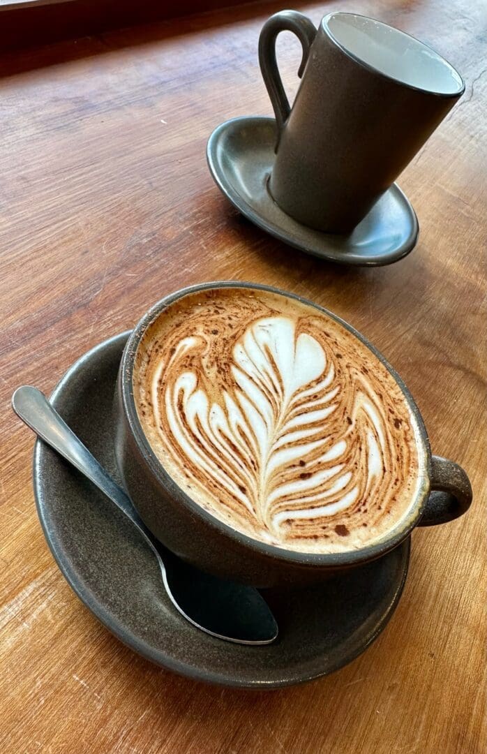 A cup of latte with intricate leaf-shaped art sits on a brown saucer with a spoon, on a wooden table. In the background, an empty matching cup hints at cozy moments found in restaurants in Frederick MD, all warmly lit by natural light.