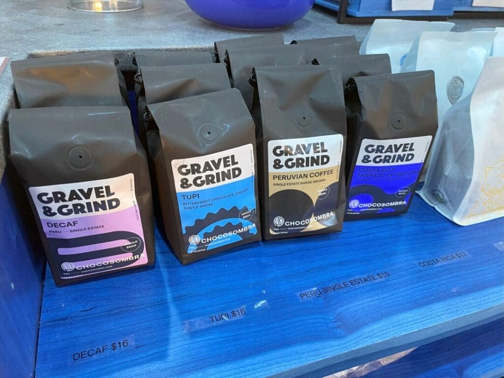 Four Black Bags of Gravel  Grind Coffee Are Displayed on a Bright Blue Shelf in a Coffee Shop Frederick Md Each Bag is Labeled Decaf Tupi Peruvian Coffee and Coopedota Rl Partial Prices and More Coffee Bags Are Visible on Either Side