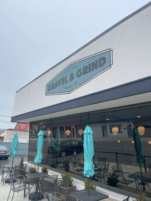 A modern cafe exterior, part of the vibrant restaurants in Frederick MD, features a sign reading Gravel & Grind above large windows. Closed teal umbrellas shade outdoor tables, with reflections of nearby buildings visible on a cloudy day.