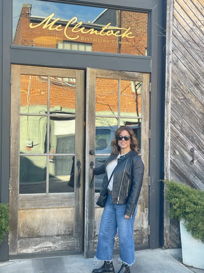 a Woman in Sunglasses a Black Leather Jacket and Blue Jeans Stands Smiling in Front of Double Glass Doors Labeled Mcclintock Distilling Company One of the Highlights Near Hotels in Downtown Frederick Md She Holds a Black Bag Amidst Potted Plants Outside the Rustic Building