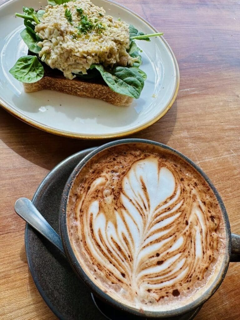 a Cup of Cappuccino with Leaf Shaped Latte Art Sits Beside an Open Faced Sandwich Topped with Spinach and a Creamy Spread All on a Wooden Table at a Cozy Coffee Shop in Frederick Md