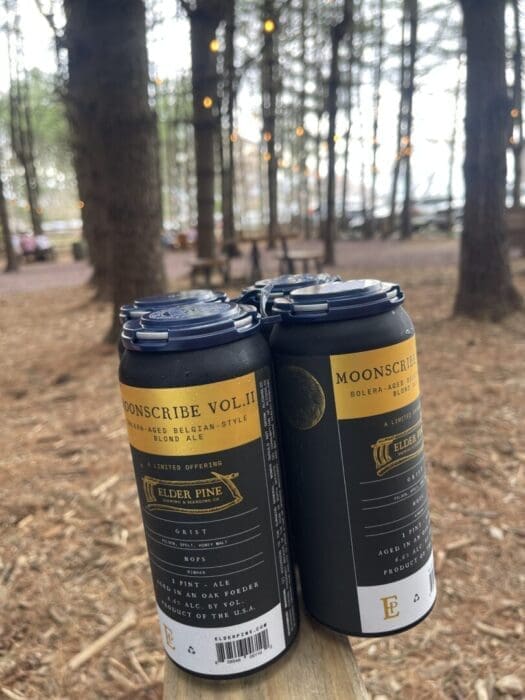 Four black and gold cans of “Moonscribe Vol. II” elder pine ale from one of the best breweries in Frederick MD sit on a wooden surface outdoors, surrounded by tall trees and warm string lights for a cozy forest vibe with blurred benches and tables.