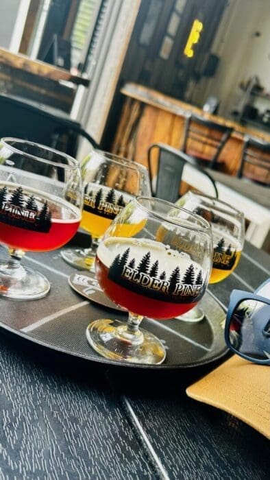 A close-up of a tray with four stemmed glasses of assorted beer at one of the best breweries in Frederick MD, each bearing the “Elder Pine” logo. The rustic, modern brewery features wood and metal décor with drinks ranging from red to amber and yellow.