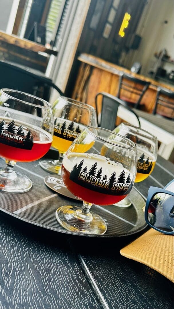 A close-up of a tray with four stemmed glasses of assorted beer at one of the best breweries in Frederick MD, each bearing the “Elder Pine” logo. The rustic, modern brewery features wood and metal décor with drinks ranging from red to amber and yellow.
