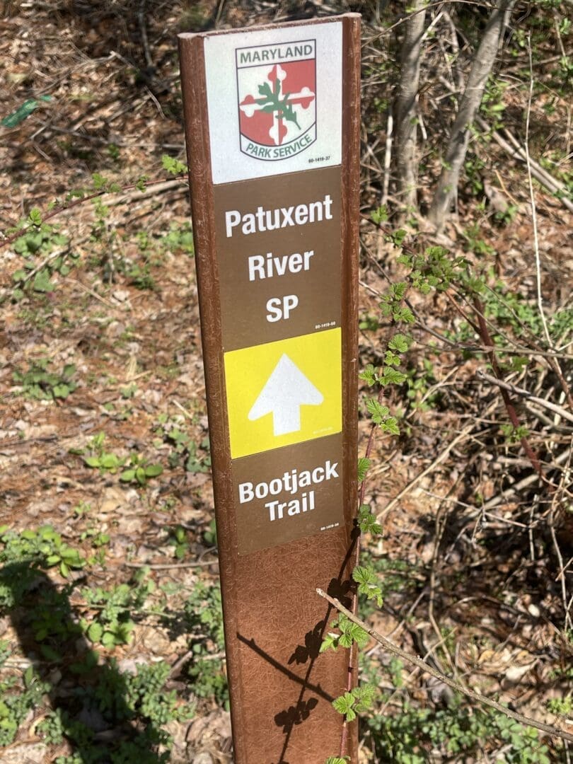 A brown trail marker post with the Maryland Park Service logo reads “Patuxent River SP” and “Bootjack Trail” with a yellow arrow. Surrounded by forest branches and leaves, it's perfect for those exploring bike trails near Frederick MD.