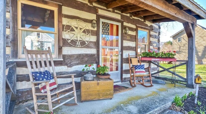 a Rustic Log Cabin Porch in Airbnb Frederick Md Features Two Rocking Chairs with American Flag Themed Cushions an Old Wooden Chest Potted Flowers and a Table with Red Geraniums Decorate the Space While a Star Decor and Flag Hang by the Door