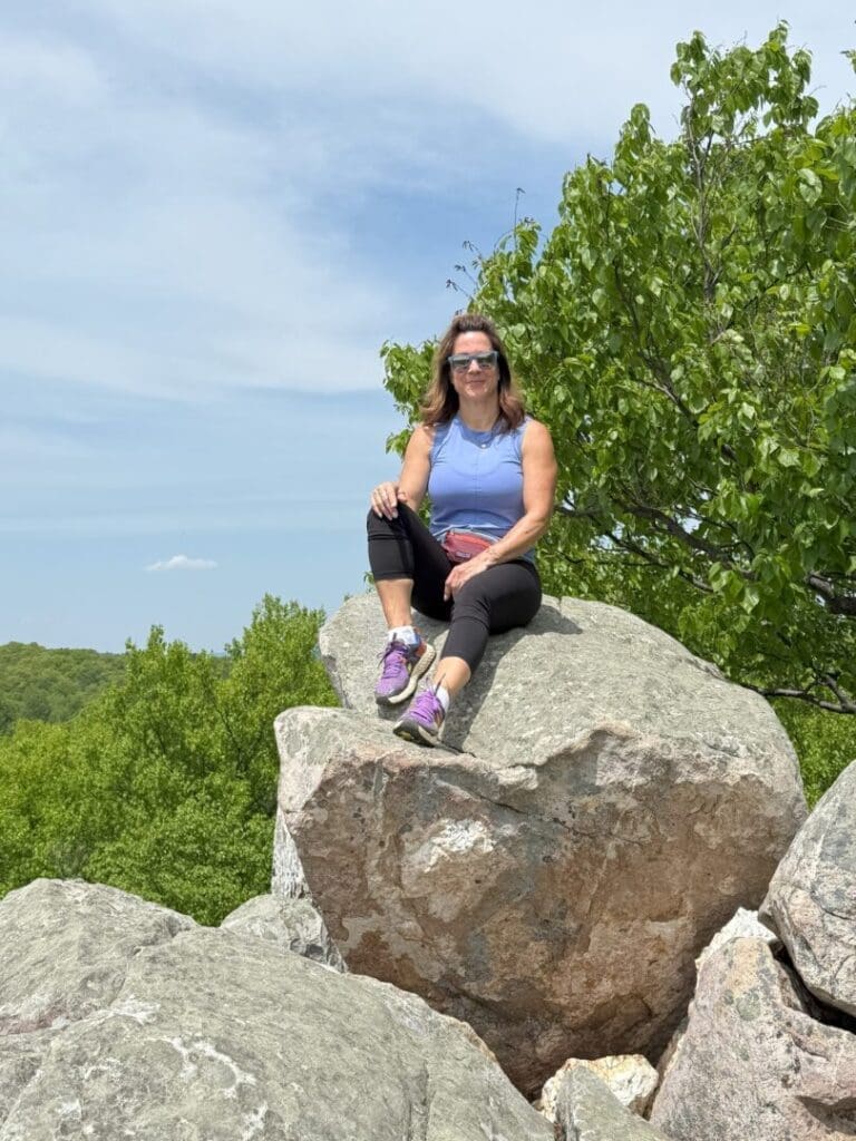 A woman with long brown hair, sunglasses, and a purple tank top sits on a large rock at Cunningham Falls State Park, surrounded by green trees under a blue sky. Smiling in black leggings, purple sneakers, and a pink waist bag, she enjoys the sunny outdoor setting.