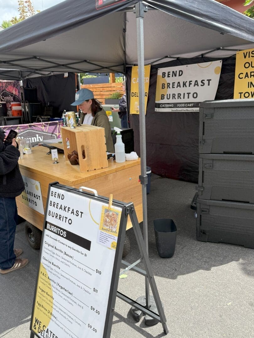 a Person Wearing a Cap Serves Customers from a Wooden Food Cart Under a Black Canopy the Sign Reads Bend Breakfast Burrito a Menu Detailing Burrito Options is Visible and a Large Black Container Stands Nearby on the Street