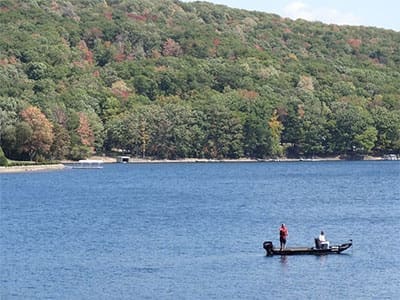 Two people are fishing from a small motorboat on a calm blue lake at Cunningham Falls State Park. Dense green forest with hints of autumn colors covers the hillside in the background, and a shoreline is faintly visible across the water.