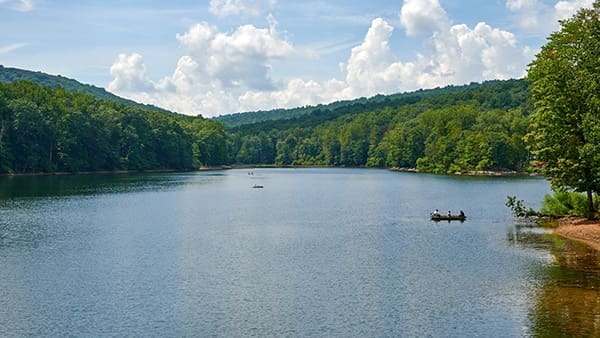 A calm lake at Cunningham Falls State Park is surrounded by green, tree-covered hills under a partly cloudy sky. Two small boats with people fishing drift on the water, while lush foliage lines the visible shoreline to the right.