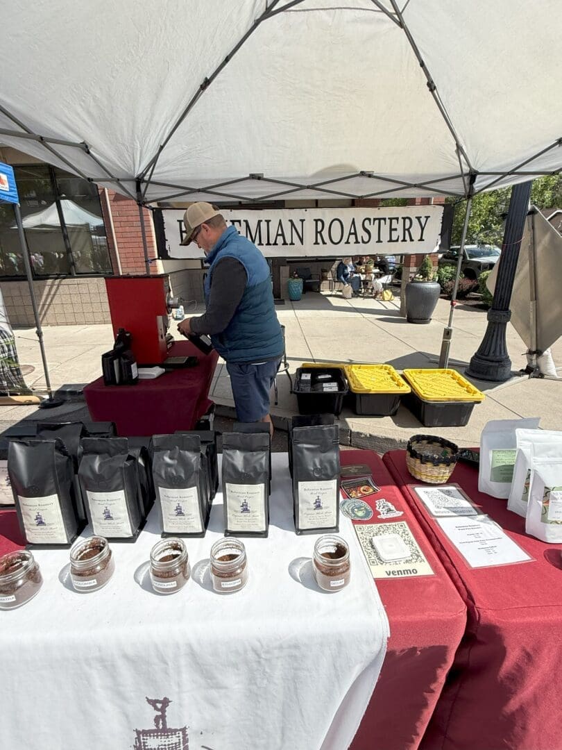 a Man Prepares Coffee Behind a Table Covered with Maroon Cloth at an Outdoor Market Stall Labeled Bohemian Roastery Various Coffee Bags Sample Jars Menus and Payment Signs Are Displayed on the Table Under a White Canopy
