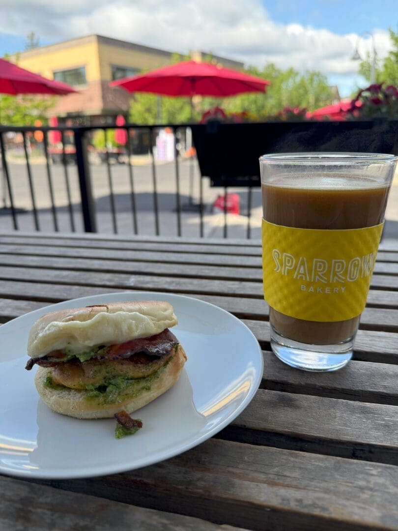 a Breakfast Sandwich with Bacon Egg and Green Arugula Sauce Sits on a White Plate Next to a Glass of Iced Coffee Labeled Sparrow Bakery on a Wooden Outdoor Table the Background Shows Red Umbrellas Trees and a Building Under a Partly Cloudy Sky