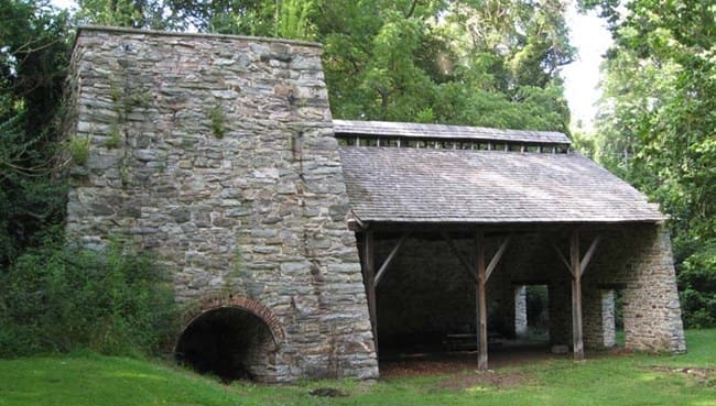 A historic stone furnace stands amid green grass and trees at Cunningham Falls State Park. The structure features a large arched opening in front, a tall stone chimney on the left, and a wooden roof supported by posts. Sunlight filters through the trees.