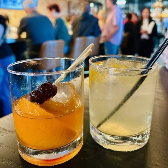 Two Cocktails in Short Glasses Sit on a Black Table at a Lively Spot Near a Coffee Shop in Frederick Md One Drink Features an Orange Slice and Cherries the Other is Pale Yellow with Ice and a Black Straw Blurred People and a Busy Bar Fill the Background