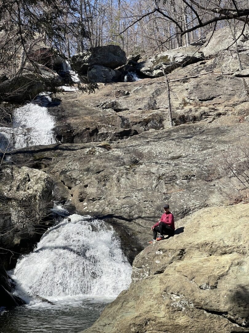 A person in a red jacket sits on a large rock at Cunningham Falls State Park, facing a small waterfall cascading over rugged terrain. Leafless trees and scattered boulders surround the scene, suggesting early spring or late fall.
