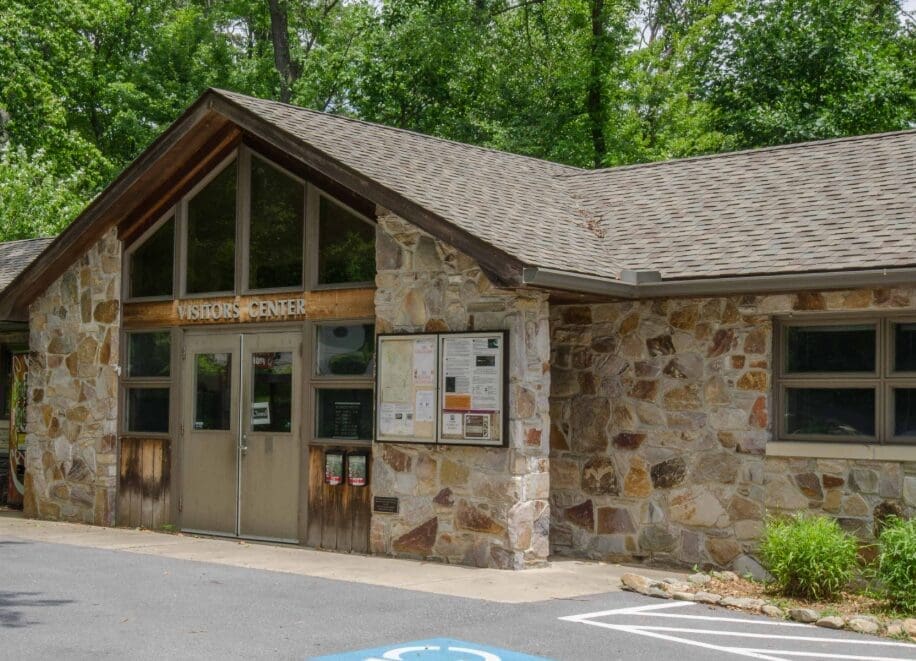A stone visitor center building with large windows and a triangular roof sits among green trees at Cunningham Falls State Park. A bulletin board and informational signs are near the door, with a paved area and part of a blue handicap parking space in front.