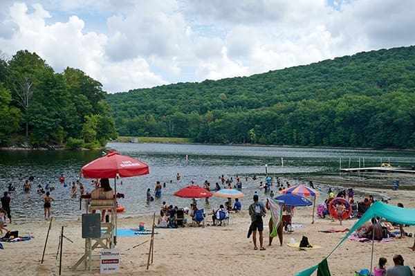 A sandy beach at Cunningham Falls State Park is crowded with people swimming, sunbathing, and relaxing under colorful umbrellas. A lifeguard watches from under a red umbrella as lush green trees and hills surround the lake under a partly cloudy sky.