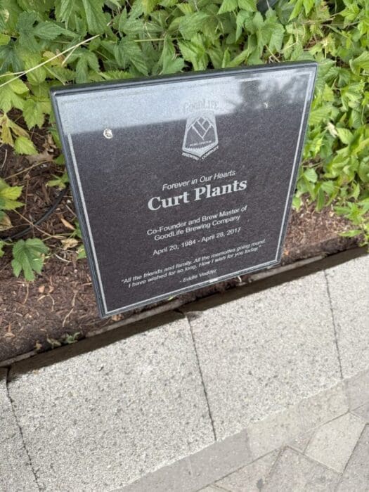 a Memorial Plaque Surrounded by Green Leafy Plants the Plaque Reads Forever in Our Hearts Curt Plants Co Founder and Brew Master of Goodlife Brewing Company April 20 1984   April 28 2017 the Plaque Includes a Quote and It Rests Beside a Concrete Sidewalk