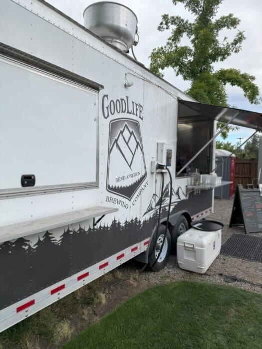 a White Food Truck Labeled Goodlife Brewing Company Bend Oregon Features a Mountain Logo and Tree Silhouettes the Side Window is Open with Hoses and Equipment Attached a White Cooler Sits Nearby and a Menu Board is Visible in the Background