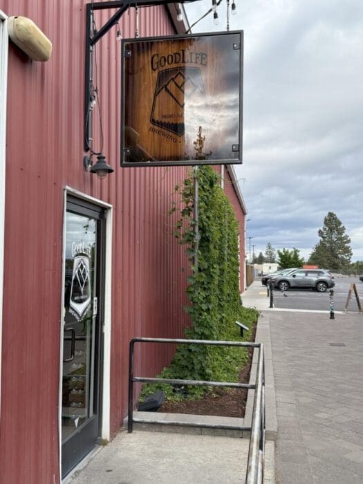 a Red Metal Building with a Glass Door and a Wooden Sign Reading Goodlife Brewing Company Green Climbing Plants Grow Up the Side and the Sidewalk Leads to a Mostly Empty Parking Lot Under a Cloudy Sky