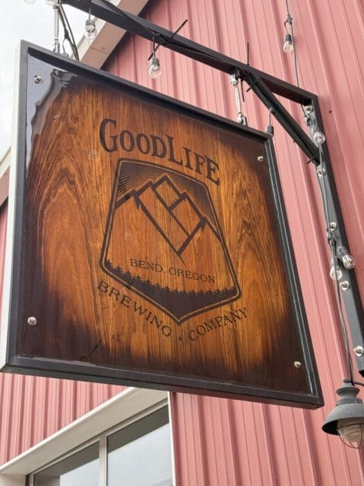 A wooden sign for GoodLife Brewing Company, one of the top breweries in Bend, Oregon, hangs outdoors on a metal frame with string lights, in front of a red building with vertical siding and a mountain logo.