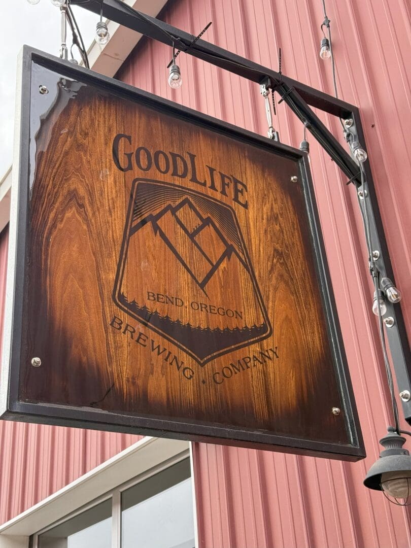 a Wooden Sign for Goodlife Brewing Company One of the Top Breweries in Bend Oregon Hangs Outdoors on a Metal Frame with String Lights in Front of a Red Building with Vertical Siding and a Mountain Logo
